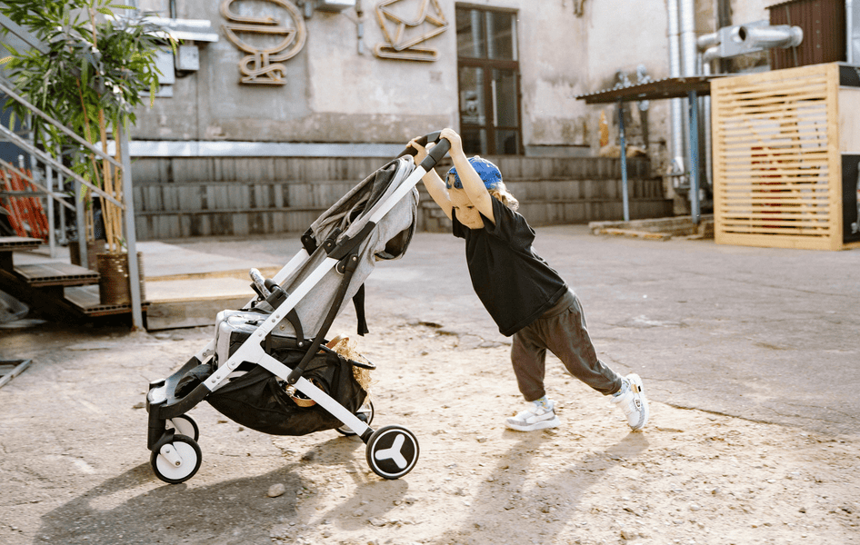 Ein kleiner Junge mit blauer Kappe schiebt spielerisch einen leichten, grauen Buggy auf einem Hof.