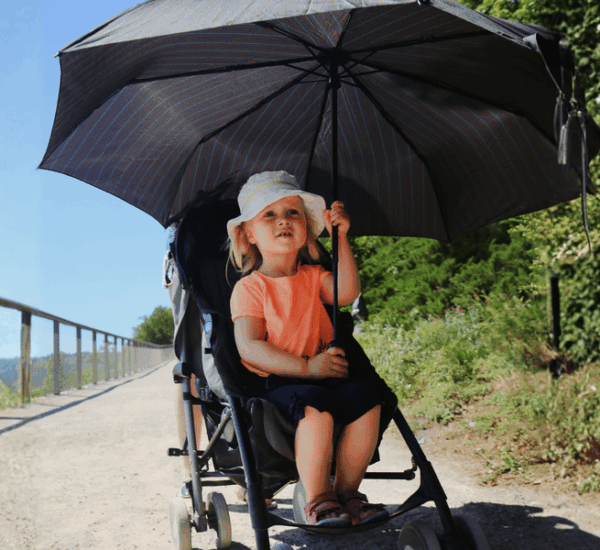 Ein kleines Mädchen sitzt im Kinderwagen unter einem großen, schattenspendenden Schirm auf einem sonnigen Weg.