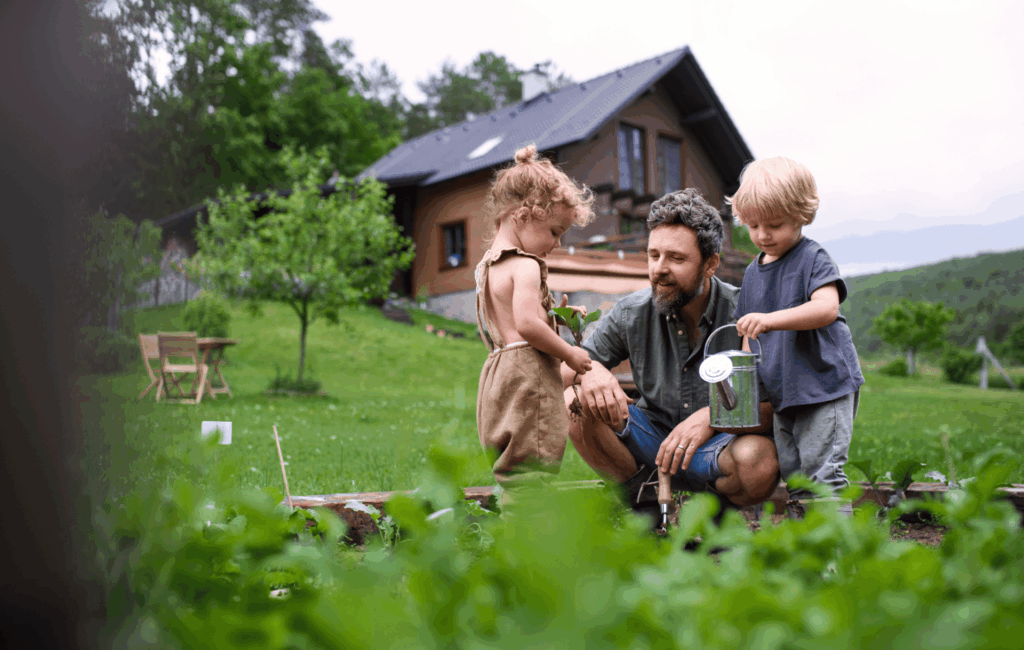 Ein Vater kniet mit seinen zwei kleinen Kindern am Holzrahmen eines Gemüsebeets hinterm Haus und hilft ihnen beim Gießen mit einer kleinen Zink-Gießkanne.